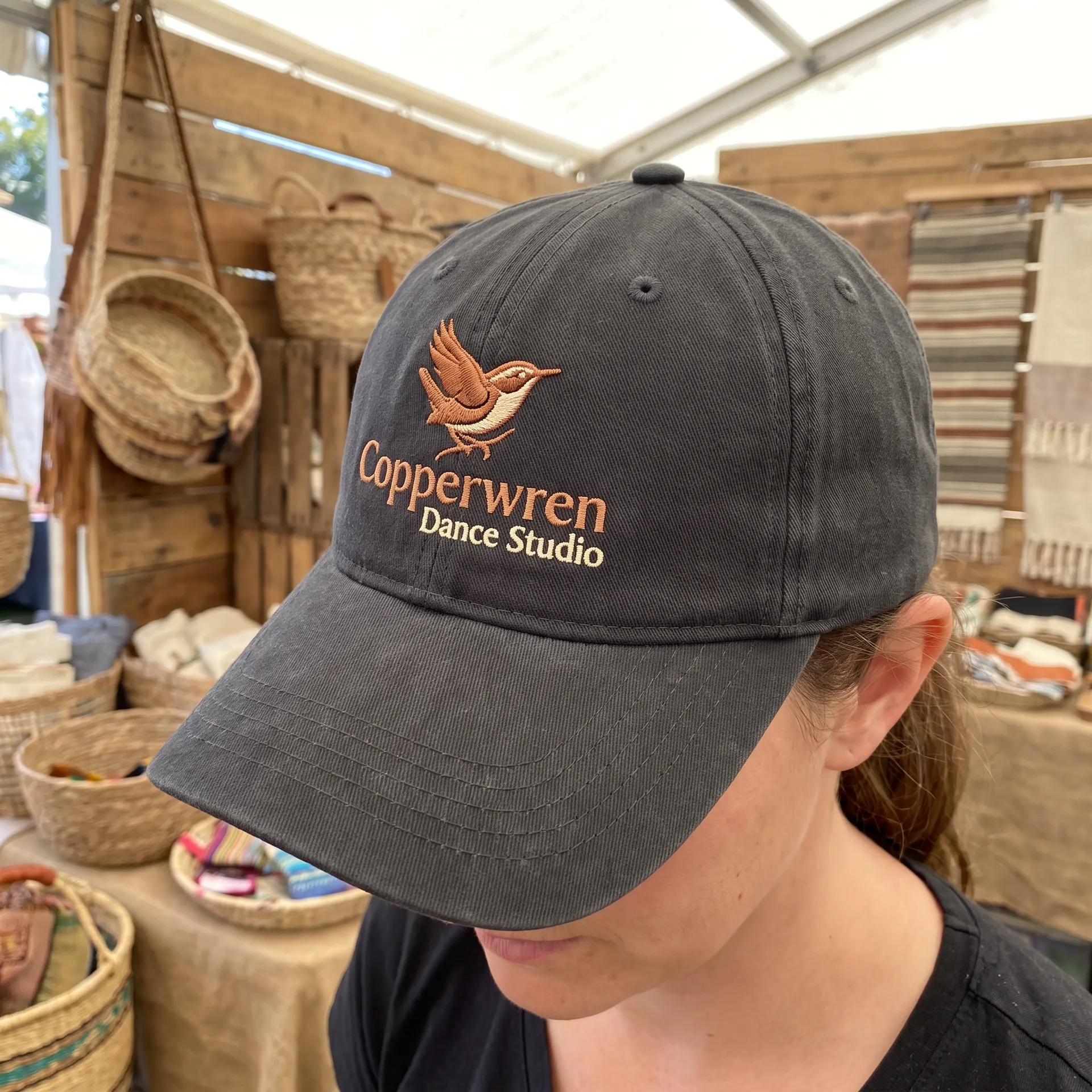 Structured six-panel cap with a front logo embroidered in raised thread, front logo, shown with a sample design: worn by a vendor at an outdoor market stall, seen from the side with the face turned away